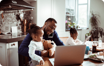 Man on cell phne sitting at kithen table with two children using laptop